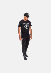 Mustat graafiset t-paidat, joissa on Raiders-logo, mustat collegehousut, joissa logo reiden päällä, ja musta lippalakki logolla. Rentouttava malli, puuvillamateriaali.