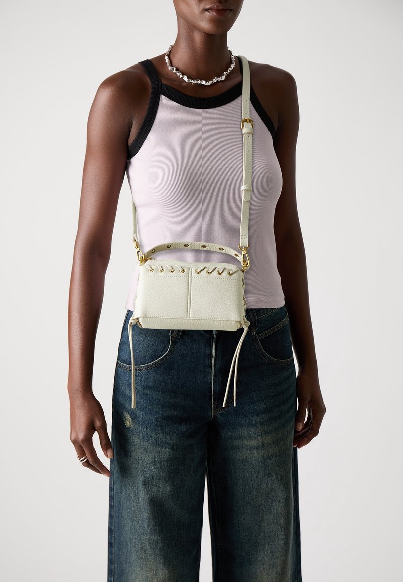 Femme portant un débardeur violet clair et un jean large foncé, portant un petit sac bandoulière écru avec des ferrures dorées et des détails en dentelle.