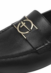 Svarta läderloafers med en texturerad yta och en gyllene cirkulär logotypaccent på ovandelen. Stickad detaljering längs sulan.