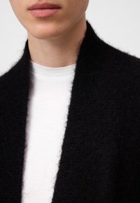 Svart ullcardigan med en strukturerad finish, med en bred öppen krage, lagrad över en vit t-shirt i bomull. Subtil kontrast i färger.