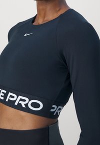 Lähikuva vartalosta, jossa on musta pitkähihaisen crop-topin, jossa on valkoinen "NIKE PRO" elastinen vyö ja pieni valkoinen Nike-kohouhlogo rinnassa.