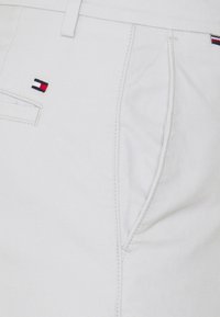 Chinos de algodón beige claro con una textura suave, que presentan un pequeño logotipo bordado y bolsillos laterales. Diseño ajustado con costuras visibles.