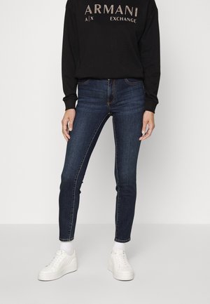 Osoba oblečená v tmavě modrých úzkých džínách, černé mikině Armani Exchange, bílých ponožkách a bílých platformových teniskách, stojící před jednobarevným pozadím.