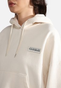 Ljusbeige hoodie i bomull, med en framfickan (kangaroo pocket), dragsko på huvan och en rektangulär logotyglapp på bröstet.