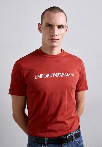 Jaunas vyras su trumpais plaukais, apsirengęs raudona Emporio Armani marškinėliais ir mėlynomis džinsinėmis kelnėmis, stovi prieš šviesiai pilką vientisą sieną.