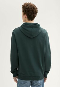 Ciemnozielony sweatshirt z kapturem, ściągaczami na rękawach i u dołu. Gładka tekstura o prostym wzorze. Widok z tyłu, noszony na jasnoniebieskich dżinsach.