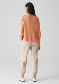 Lichtgewicht, perzikkleurig sweater met een geribde onderrand en driekwartmouwen, gecombineerd met beige joggingbroek en witte sneakers.