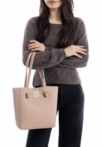 Bolso tote de imitación piel beige con dos asas anudadas, que presenta un emblema dorado en la parte frontal y un diseño estructurado.