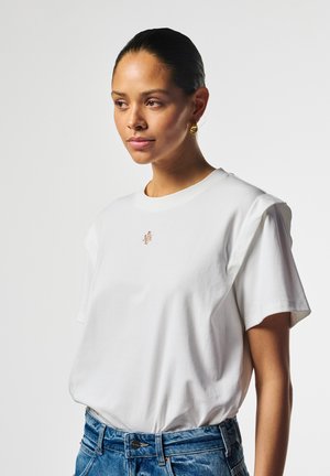 Vrouw met donker haar die gouden hoepeloorbellen draagt, een wit T-shirt met een klein logo aan de voorkant en een blauwe spijkerbroek tegen een egale lichte achtergrond.