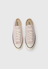 Zapatillas de lona rosa claro con puntas de goma crema, rayas negras y cordones beige. Diseño clásico de caña baja con textura suave y ojales plateados.