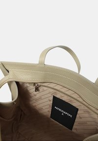 Interno della borsa tote in pelle tan con una tasca con zip, fodera in tessuto a fantasia e un'etichetta logo nera con testo bianco. Texture liscia, forma strutturata.