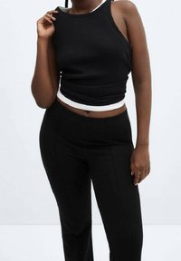 Femme portant un crop top noir sans manches avec un ourlet blanc et un pantalon noir ajusté, debout devant un fond clair uni.