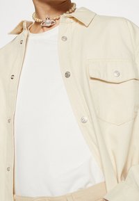 Light beige button-up shirt with silver hardware, worn over a white t-shirt. Features a chest pocket and a pearl choker with a silver accent.