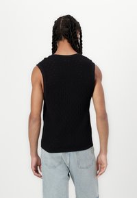 Sort ærmeløs strikvest med diamantmønster og hulmønstrede detaljer, med rund halsudskæring og ribbet afslutning. Båret med lyseblå jeans.