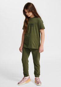 Oliwkowa bawełniana koszulka z krótkim rękawem i nadrukowanym tekstem, w połączeniu z dopasowanymi spodniami dresowymi i jasnoróżowymi sneakersami sportowymi.