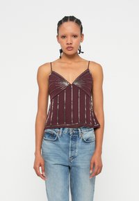 Burgundska kamizola s tankimi naramnicami, ki ima vertikalne kovinske okraske in oblikovano srčno dekolte, v kombinaciji s svetlimi jeans hlačami.