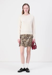Krēmveida džemperis, leoparda raksta svārki, melni loaferi, baltas zeķes un maza sarkana rokas soma uz vienkārša baltā fona.