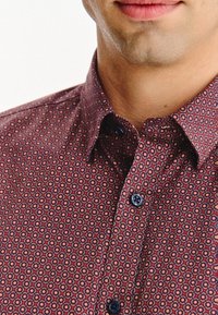 Bordowa koszula z guzikami, z geometrycznym wzorem w odcieniach czerwieni, żółci i niebieskiego. Posiada klasyczny kołnierzyk i czarne guziki.