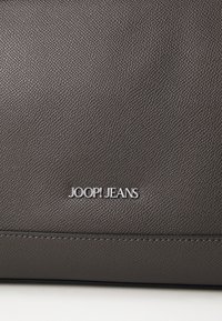 Borsa in pelle marrone con texture, caratterizzata da un logo argento "JOOP! JEANS", con un design pulito e cuciture visibili lungo la base.