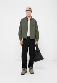 Green button-up jacket with a patch, white t-shirt, black cargo pants, beige cap with blue and brown accents, gray boots, and a black bag.