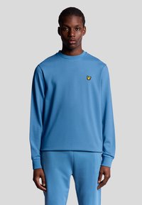 Felpa azzurro chiaro a maniche lunghe con colletto girocollo e un piccolo logo giallo sul petto; tessuto morbido e liscio.