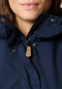 Gros plan du bas du visage et du cou d’une personne portant une veste bleu marine avec un col partiellement zippé et une tirette en cuir.