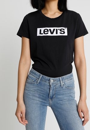 Kvinde iført sort Levi's t-shirt og lyseblå høj talje jeans står mod en ensfarvet baggrund.
