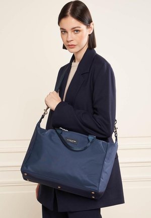 Sac en nylon bleu marine avec double poigné et une bandoulière amovible, arborant un logo à l'avant et des accents dorés sur les pieds.