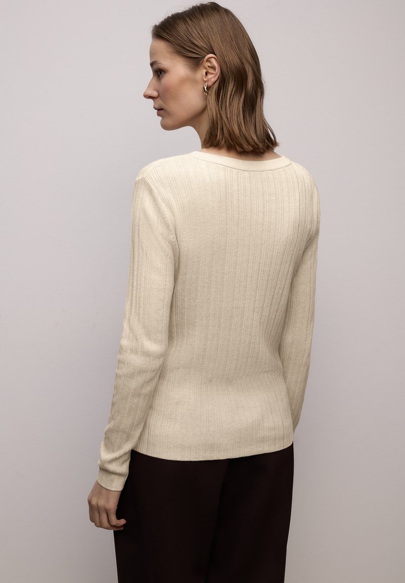 Maglione crema a coste in maglia con maniche lunghe e scollo rotondo, caratterizzato da una silhouette aderente e una texture sottile. Abbinato a pantaloni scuri.