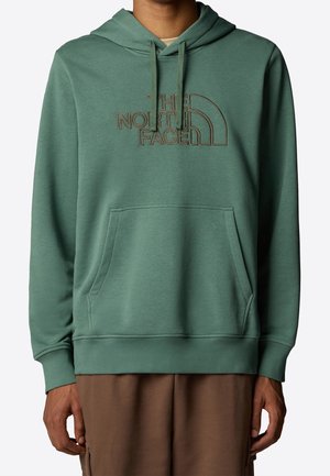 Grüner Hoodie mit Kängurutasche und auf der Brust besticktem "The North Face"-Logo, getragen mit braunen Hosen.