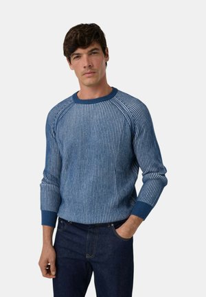 Hombre con un suéter de punto acanalado azul de manga larga y vaqueros azul oscuro, con una mano en el bolsillo, sobre un fondo claro y liso.