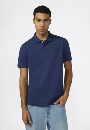 Jonge man die een marineblauw polohemd met korte mouwen en lichtblauwe jeans draagt, staand tegen een effen witte achtergrond.