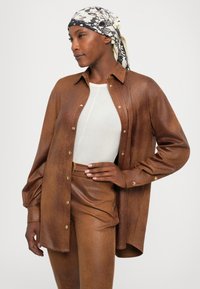 Brown faux leather shirt with a relaxed fit, buttoned front, and long sleeves. Worn over a fitted white top, accessorized with a patterned scarf.