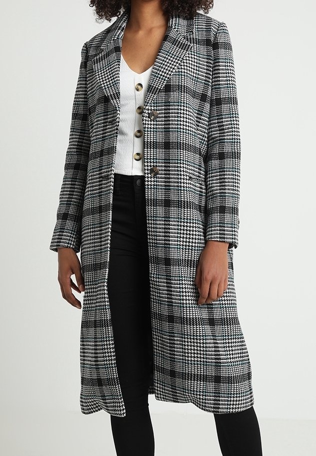 Femme portant un long manteau à carreaux noir et blanc par-dessus un haut blanc boutonné et un pantalon noir, debout, les mains détendues le long du corps.