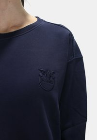 Felpa blu navy con un logo rotondo in rilievo raffigurante uccelli e il nome del marchio "PINKO" nella parte sinistra del petto. Tessuto morbido.