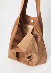 Bruine suède schoudertas met een zachte textuur, open bovenkant en een drukknoopsluiting. Het interieur heeft een ritsvak en een beige voering.