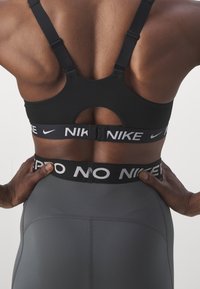 Svart treningstopp med utskjæringsdesign og justerbare stropper, med Nike-logo. Grå tights med svart midje med teksten "NO NIKE".
