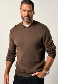 Felpa con cappuccio in tessuto a maglia marrone scuro con polsini e orlo a costine, indossata sopra una camicia bianca; uomo con capelli corti e un orologio.