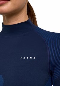 Top sportivo maniche lunghe color blu navy con collo a lupetto, caratterizzato da strisce blu testurizzate e logo bianco "FALKE" sul davanti. Tessuto liscio ed elastico.