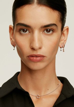 Collar de plata en forma de estrella sobre un cuello; los pendientes de la persona que lo lleva puesto son pequeños aros con charms de estrella y letras; piel suave; camisa oscura.