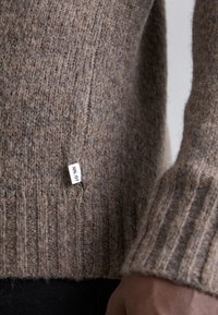 Brun ullgenser med ribbestrikkede mansjetter og kant. Har et sideetikett med "NN_07." Teksturen ser myk og strikket ut.