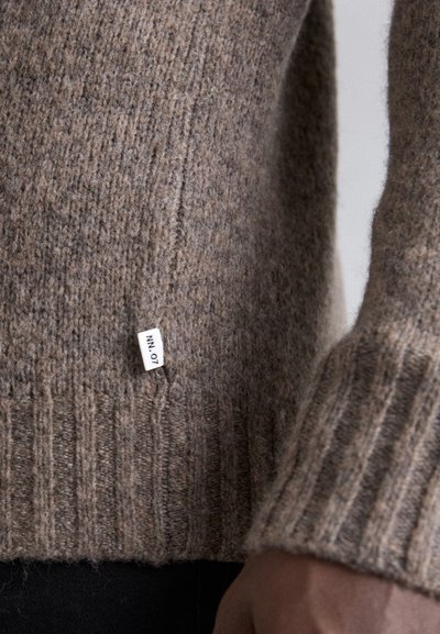 Bruine wollen trui met ribbels aan de mouwen en onderkant. Heeft een zijlabel met "NN_07." De textuur lijkt zacht en gebreid.