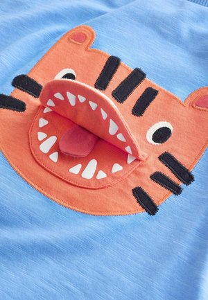 Blauer Stoff mit einem orangefarbenen Tiergesicht, dessen geöffnetes Maul eine rosa Zunge zeigt. Schwarze Streifen und weiße Zähne sind detailliert dargestellt.