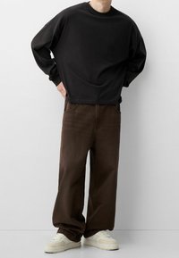Sudadera negra de manga larga, de corte relajado, combinada con vaqueros de mezclilla marrón holgados. Zapatillas de color crema completan el conjunto.