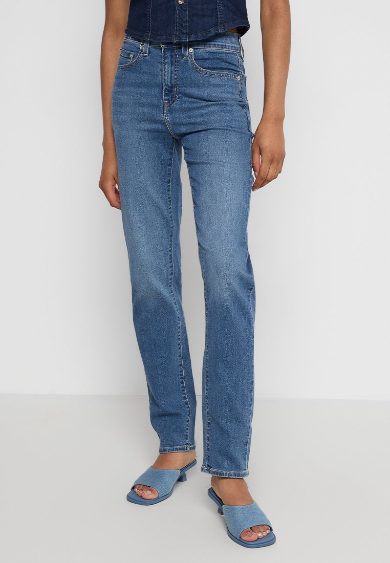 Person, die hochtaillierte gerade blaue Jeans trägt, kombiniert mit hellblauen offenen Absatzsandalen vor einem schlichten Hintergrund.