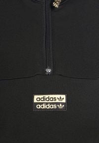 Černá zipová mikina vyrobená z elastického materiálu. Obsahuje dvoubarevnou nášivku loga Adidas na hrudi a zipové zapínání s malým táhlem.