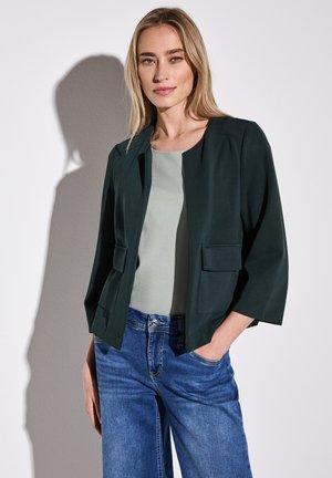 Donkergroene blazer met een ronde kraag en twee voorzakken, gecombineerd met een lichtgroene top en een blauwe wijde broek. Gladde stofstructuur.