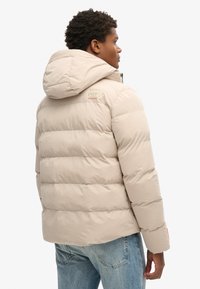 Beige pufferjakke med hætte og vandret syninger. Har logo detaljer på skulderen. Matchet med lyseblå jeans.