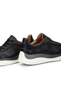 Zapatillas negras con parte superior de malla texturizada y cuero liso, suela de amortiguación blanca y acentos de costura en negro. Cordones integrados en el diseño.