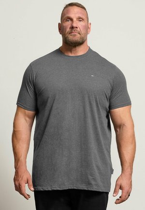 Uomo muscoloso con barba curata sta in piedi rivolto in avanti, indossa una semplice t-shirt grigio scuro a maniche corte e pantaloni neri su sfondo chiaro.